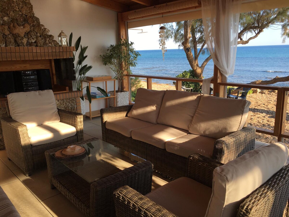 A furnished outdoor seating area includes a comfortable sofa and armchairs with soft cushions. A glass table sits in the center, while views of the sandy beach and sea can be seen through the open wall, creating a relaxed atmosphere.