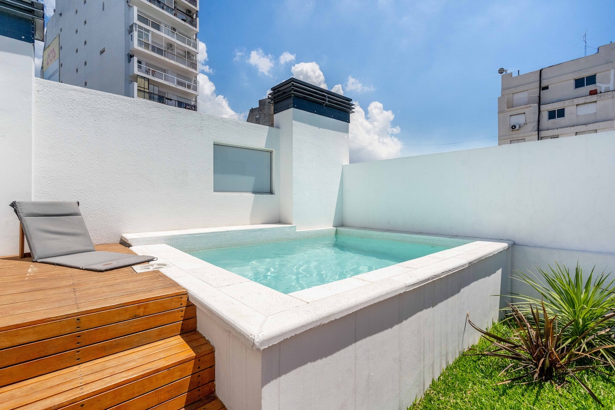 A private pool is surrounded by white walls and wooden decking, creating a serene outdoor space. Lush green grass and a desert plant are positioned near the poolside. Clear blue skies and soft clouds provide a bright backdrop to this tranquil setting.