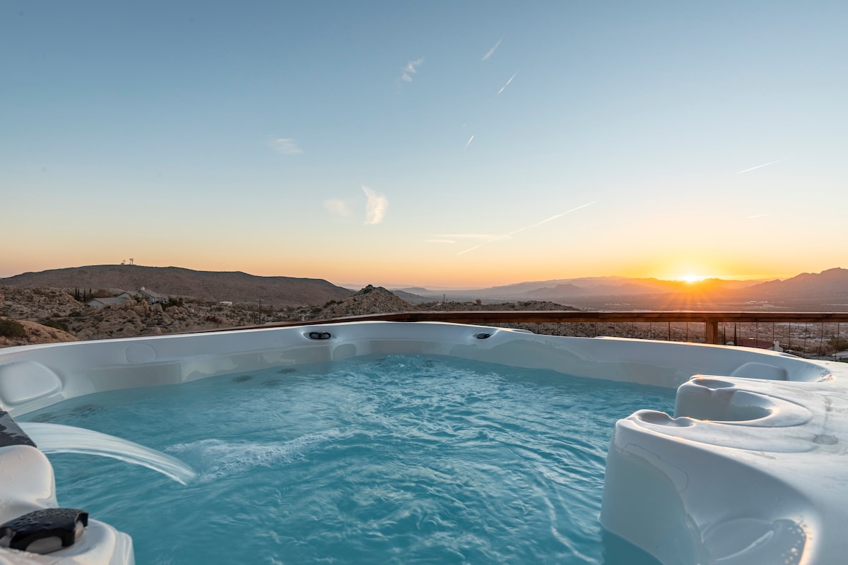 A spacious hot tub filled with clear water overlooks a stunning sunset. The surrounding landscape features rolling hills and rocky terrain, creating a serene backdrop. Sunlight casts a warm glow over the scene, highlighting the outdoor area designed for relaxation.