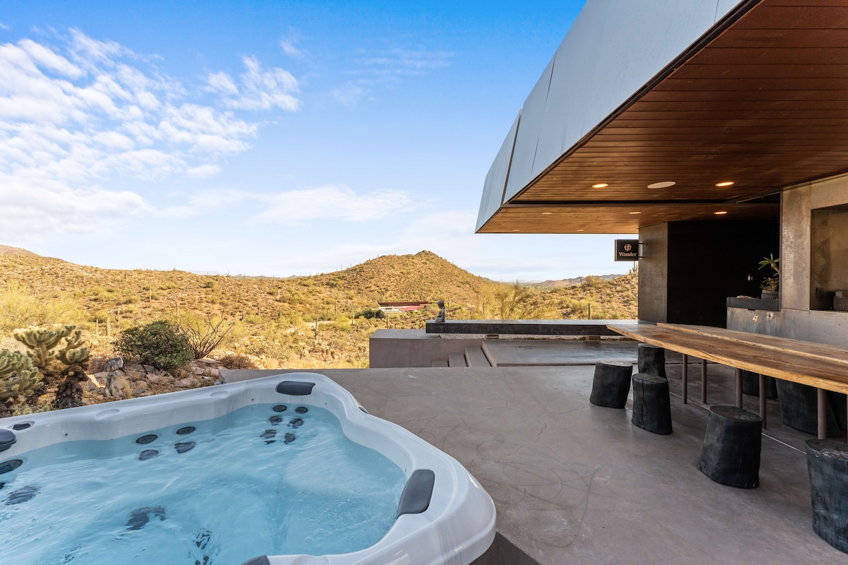 An outdoor space features a hot tub surrounded by gorgeous desert scenery and mountains. A modern dining area with a long table and black stone stools is nearby, offering inviting views of the natural landscape under a clear blue sky.