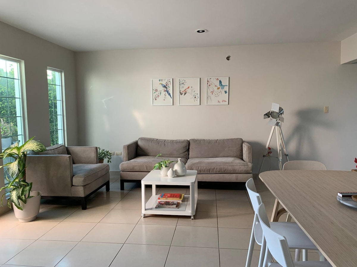 An open living area features a light-colored sectional sofa and an armchair, accompanied by a table with a few decorative items. A dining table with white chairs sits nearby. Natural light enters through large windows, illuminating the minimalist decor and artwork on the walls.