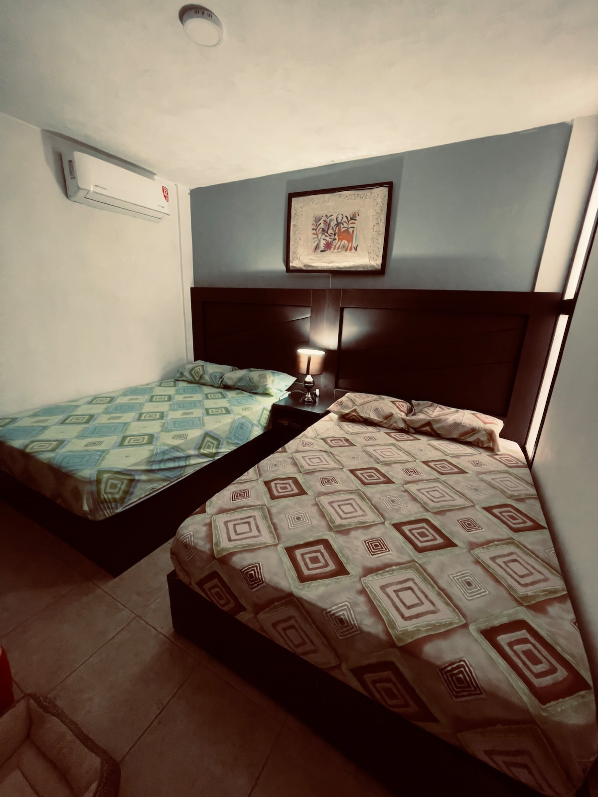 The bedroom features two spacious beds with distinct patterned bedding. A nightstand with a lamp is positioned between the beds. The walls are painted in soft blue, and an air-conditioning unit is mounted above for comfort.