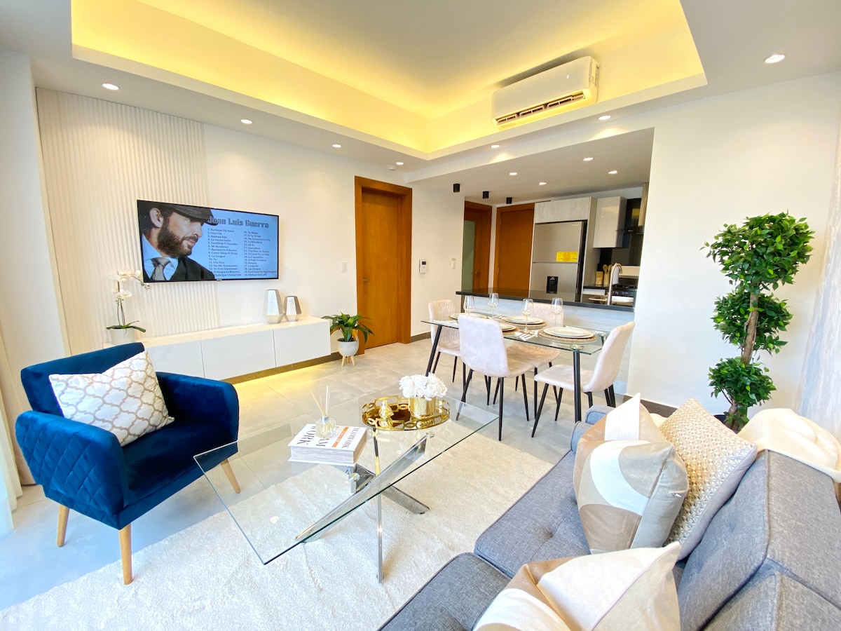 A modern living area features contrasting blue and gray seating arrangements, with a glass coffee table at the center. A wall-mounted television is positioned opposite. A dining table with white chairs is visible in the background, complemented by elegant decor, including potted plants.