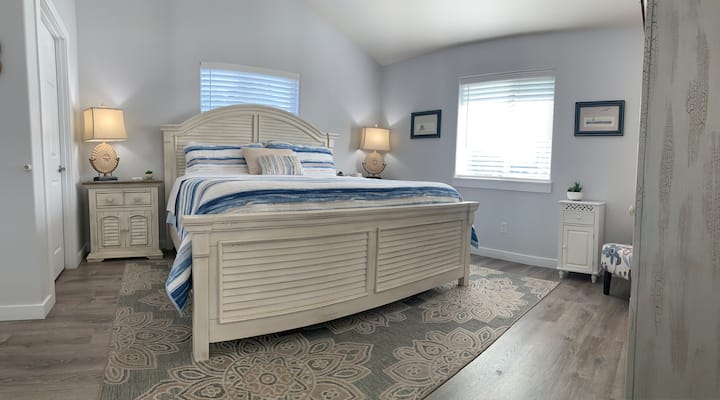Vaulted ceilings, bright windows and soft clean linens gives the primary bedroom a fresh inviting feel. Offered is a king size bed with two night stands, an armoire for unpacking and a sitting area for resting and relaxing.