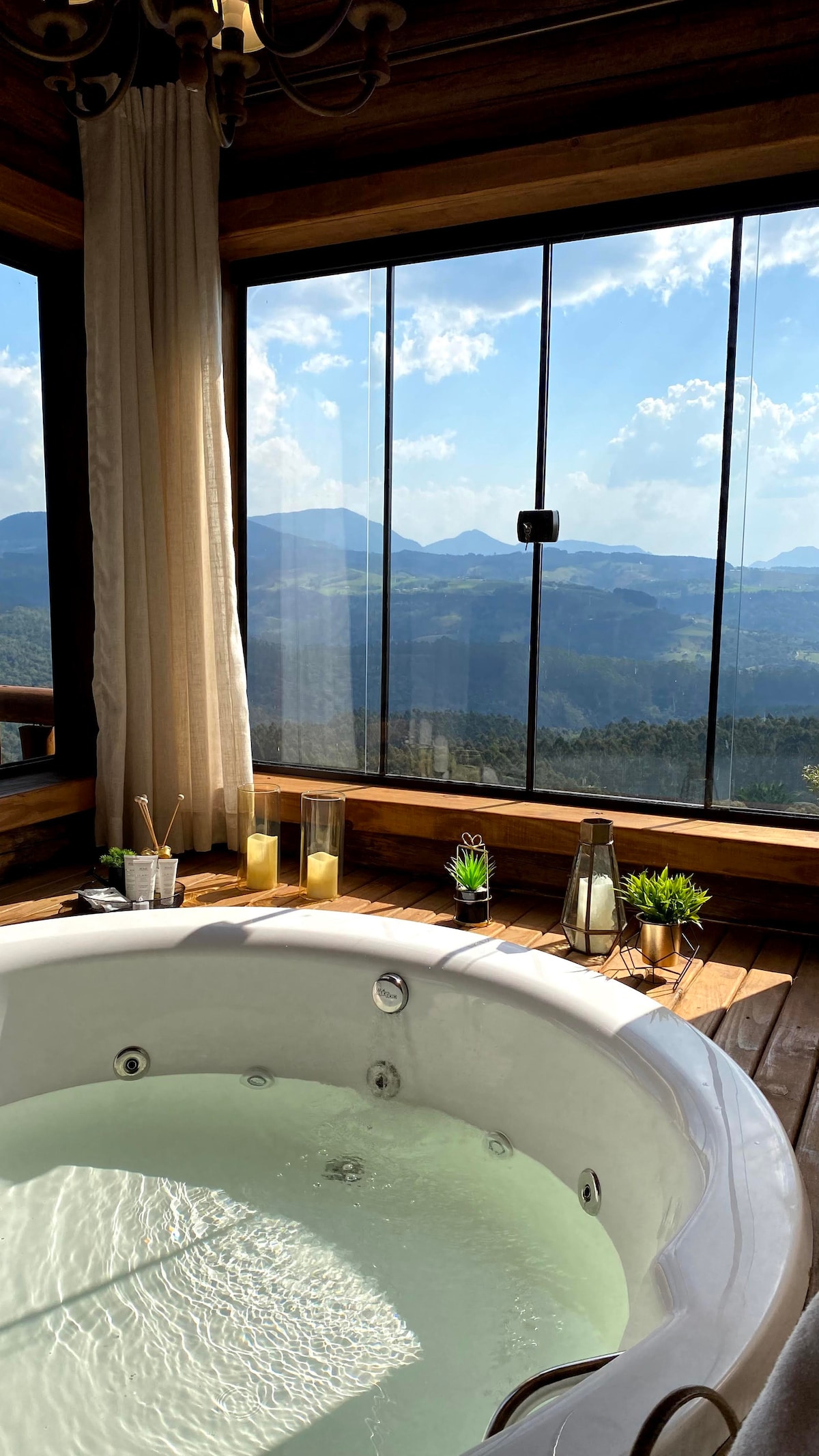 A modern hydromassage bathtub is positioned near expansive glass windows, offering an unobstructed view of the surrounding mountains and valleys. Soft natural light enters the space, and decorative elements such as candles and small plants enhance the serene atmosphere.