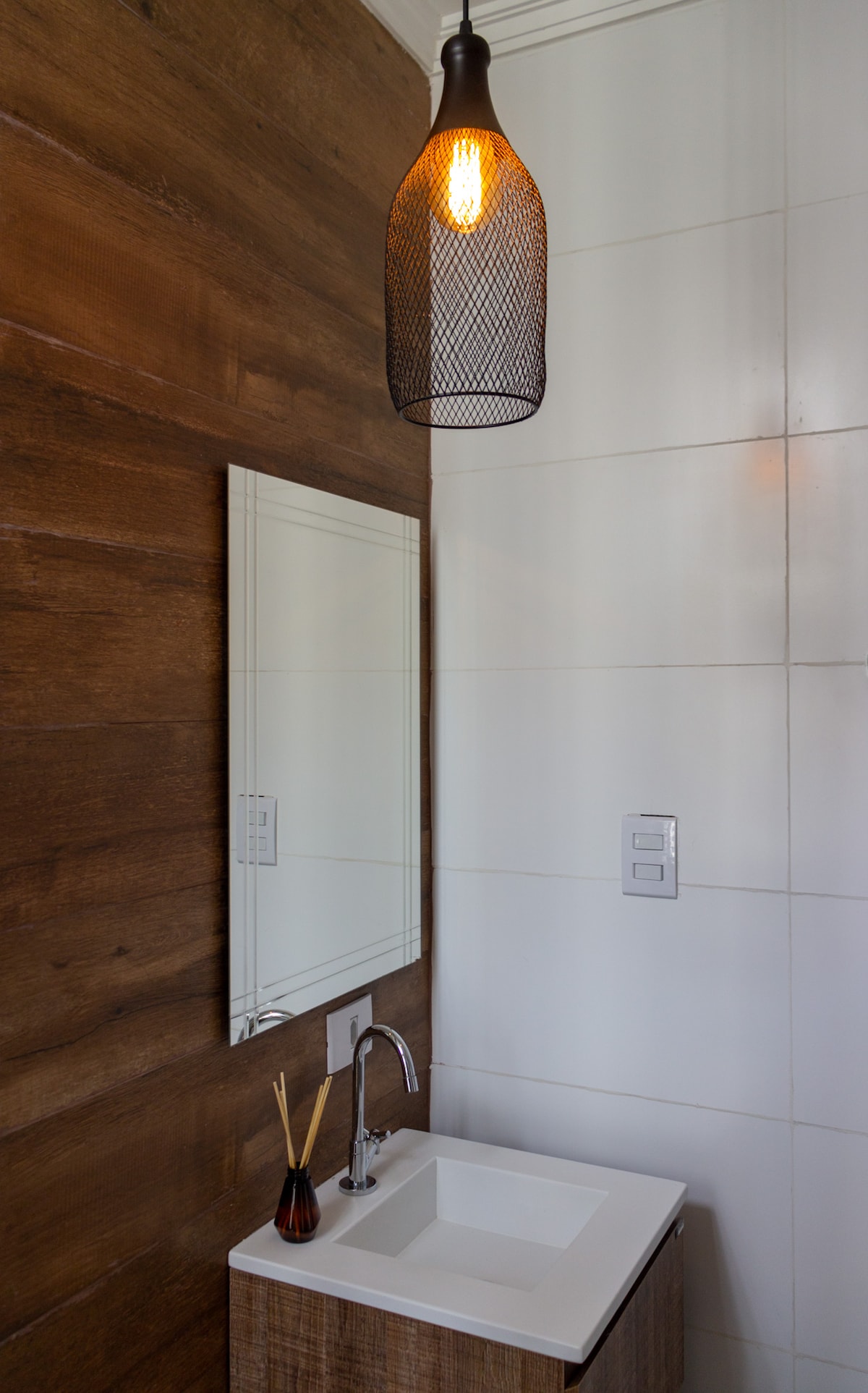 A modern bathroom features a sleek white sink set against a textured wooden wall. A stylish pendant light hangs above, providing illumination. A mirror is mounted above the sink, and a subtle decorative element rests nearby, enhancing the simple yet functional design.