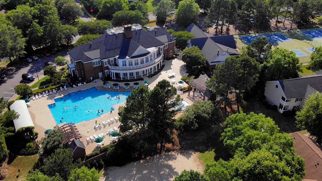 An aerial view captures the expansive resort grounds, featuring a large outdoor pool surrounded by lounge chairs. Adjacent to the pool, tennis courts and well-maintained landscaping enhance the recreational environment, offering a welcoming atmosphere for relaxation and leisure activities.