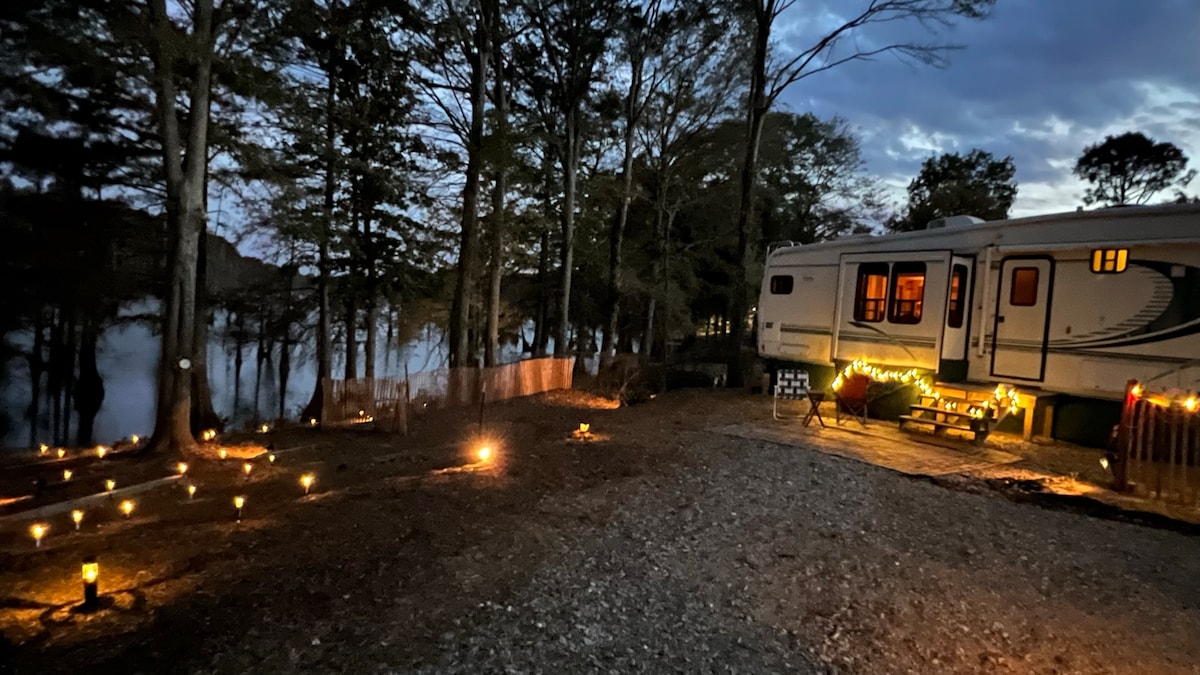The exterior of the RV is illuminated by soft candlelight, creating a serene ambiance. Trees surround the area, and reflections of the water can be seen in the background. The scene captures a peaceful evening setting, highlighting the proximity to the lake.