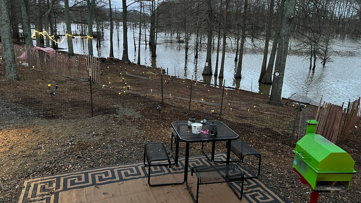 A patio area features a black metal table with two chairs, positioned beside a green grill. The setting overlooks a body of water surrounded by trees, enhancing the outdoor experience. A fenced perimeter offers added privacy, while lights are scattered along the path in the background.