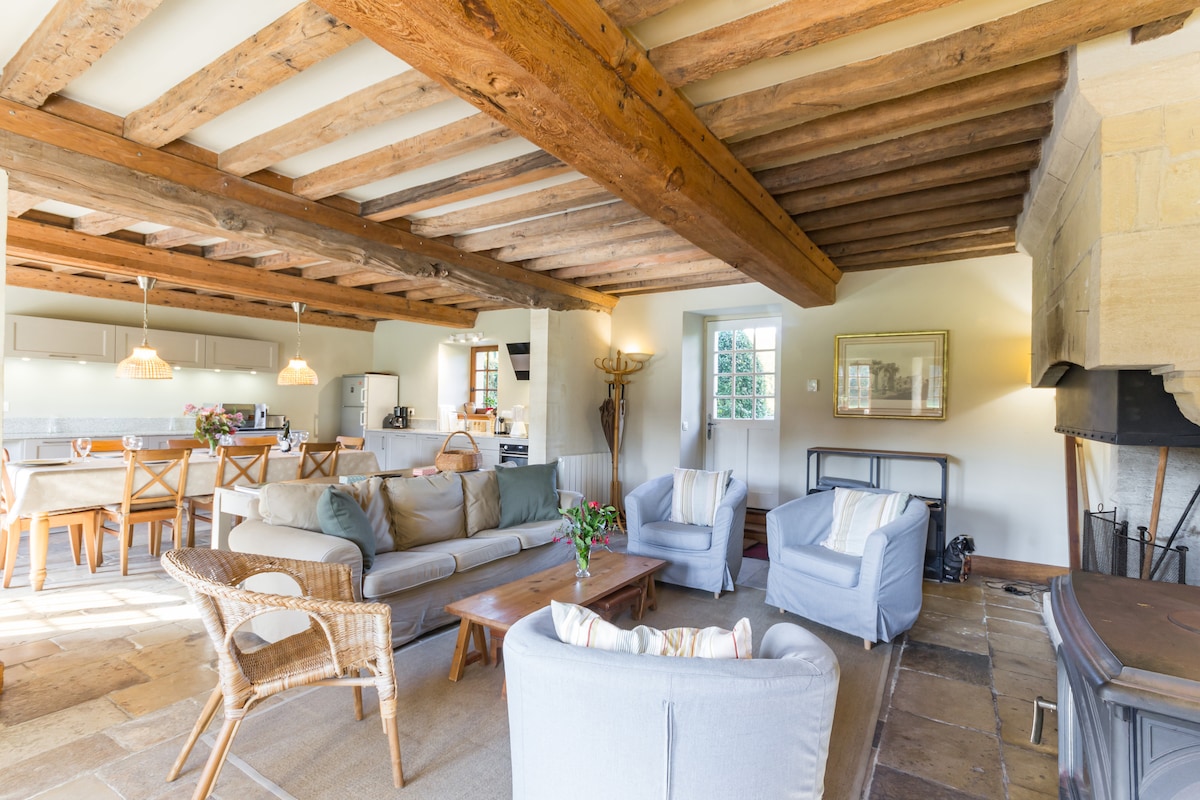 A spacious living area is characterized by wooden beams and a stone fireplace. Comfortable seating options include a light-colored sofa and two armchairs paired with a coffee table. The open design connects to a dining area that features wooden chairs and pendant lighting.