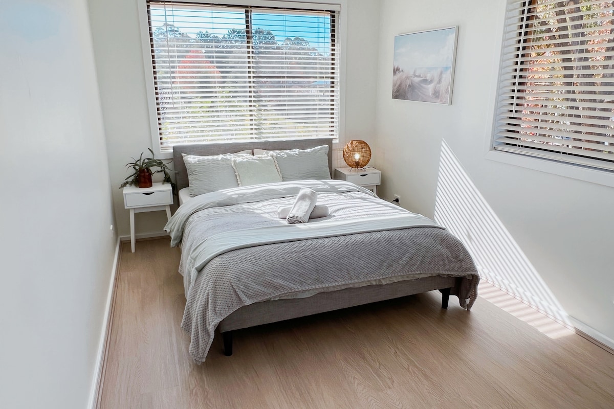 A comfortable bedroom features a spacious bed with a light grey blanket and a neatly folded towel placed on top. Natural light flows in through two windows, with wooden blinds partially opened. A small plant adorns the nightstand beside the bed.