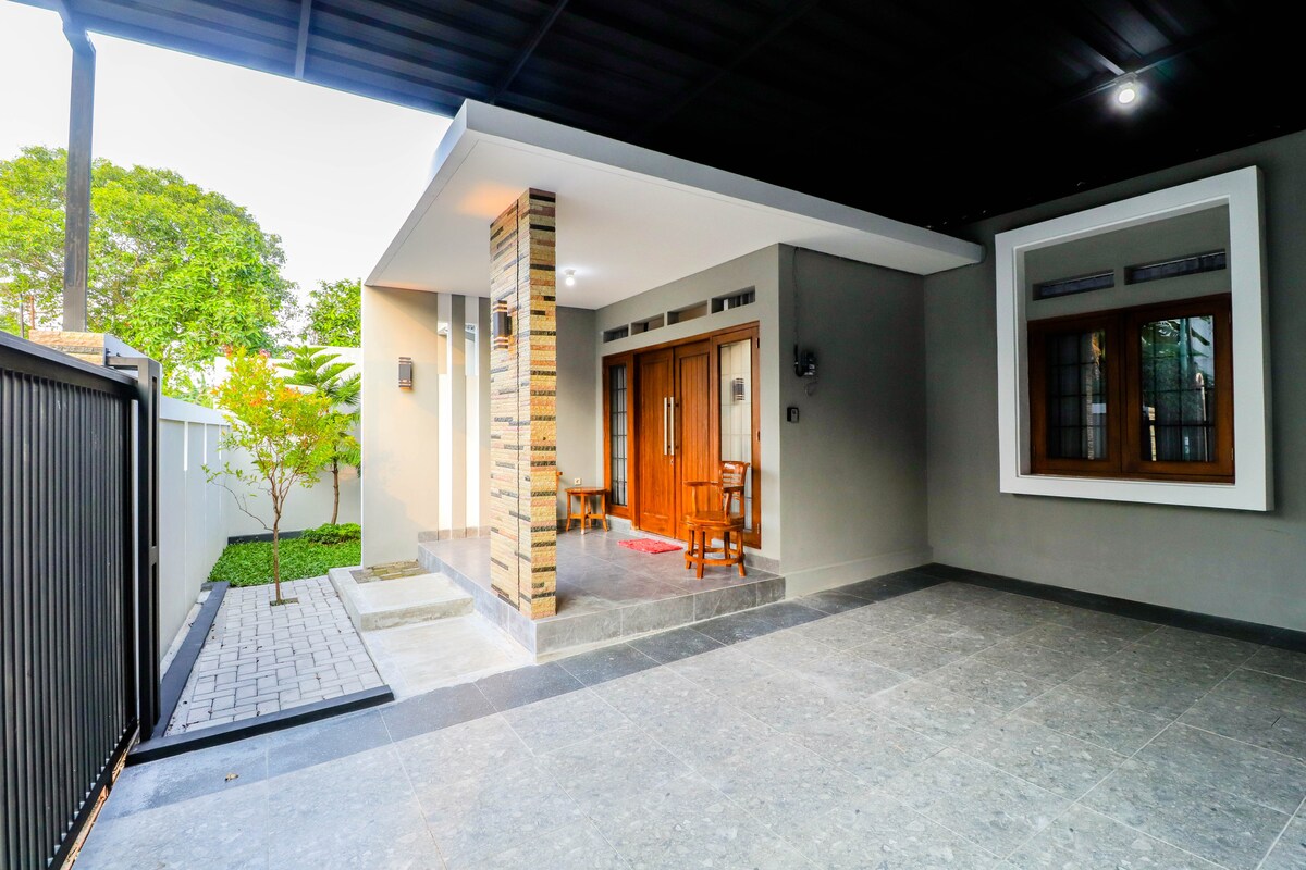 The exterior entrance features a covered porch with a stone accent wall. Two wooden chairs are placed on a tiled surface that leads to the main door. Greenery is visible in the background, adding a touch of nature to the modern design.