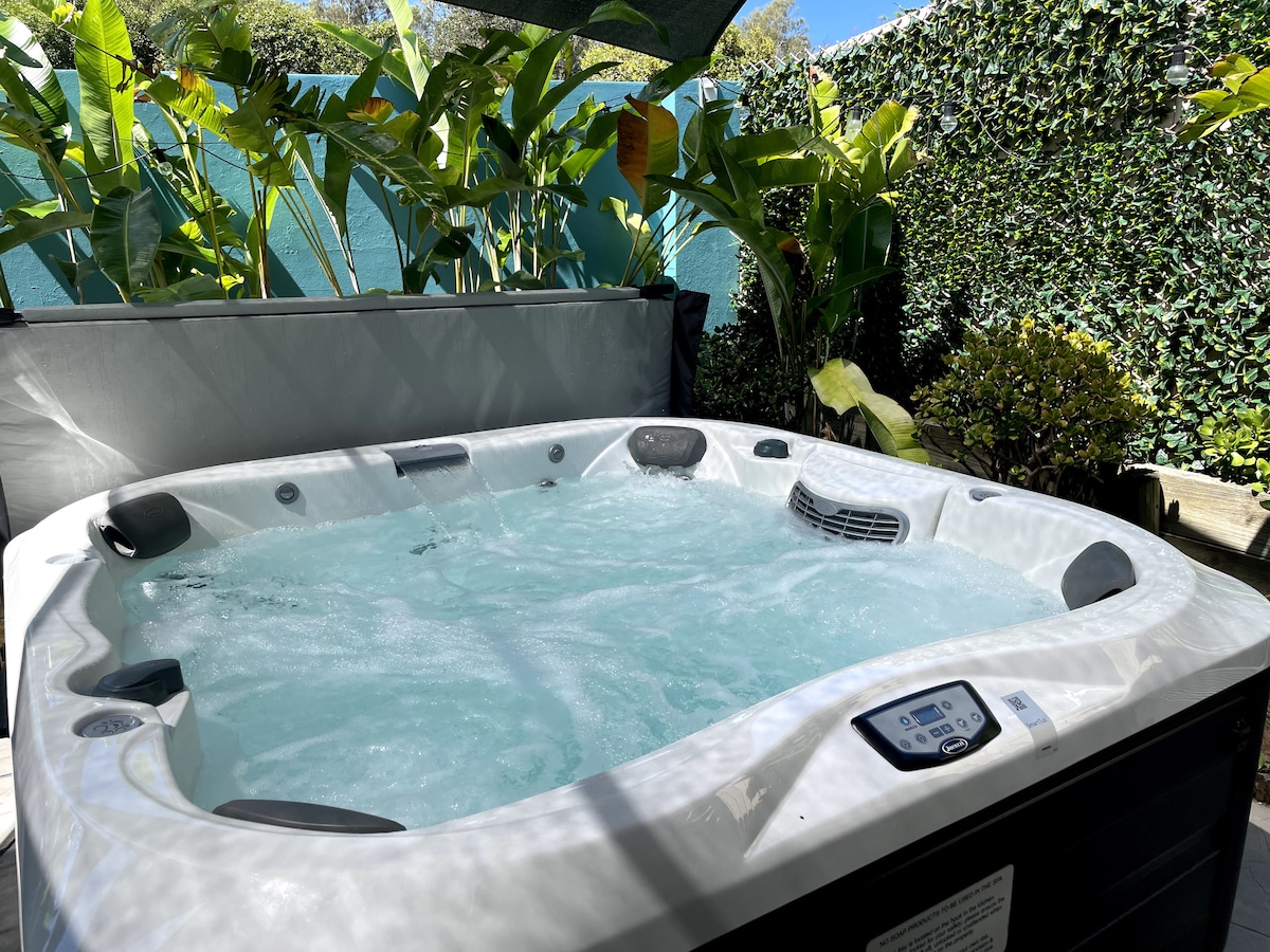 A spa is set amidst lush greenery, surrounded by tropical plants. Bubbles gently rise in the water, while the control panel is easily accessible on the side.