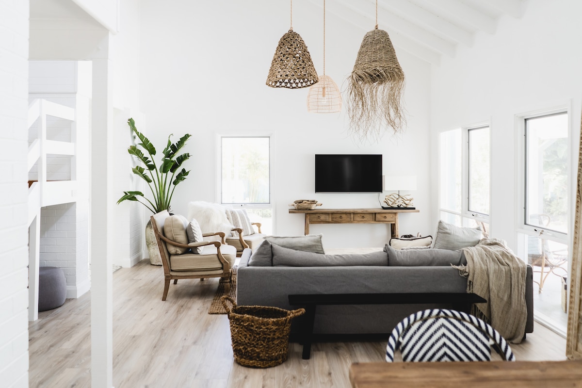 A modern living room features light wood flooring and high ceilings. A spacious grey sofa is complemented by natural woven pendant lights. Potted plants are positioned near large windows, allowing natural light to illuminate the space. A wooden side table and decorative items are arranged on a console against the wall.