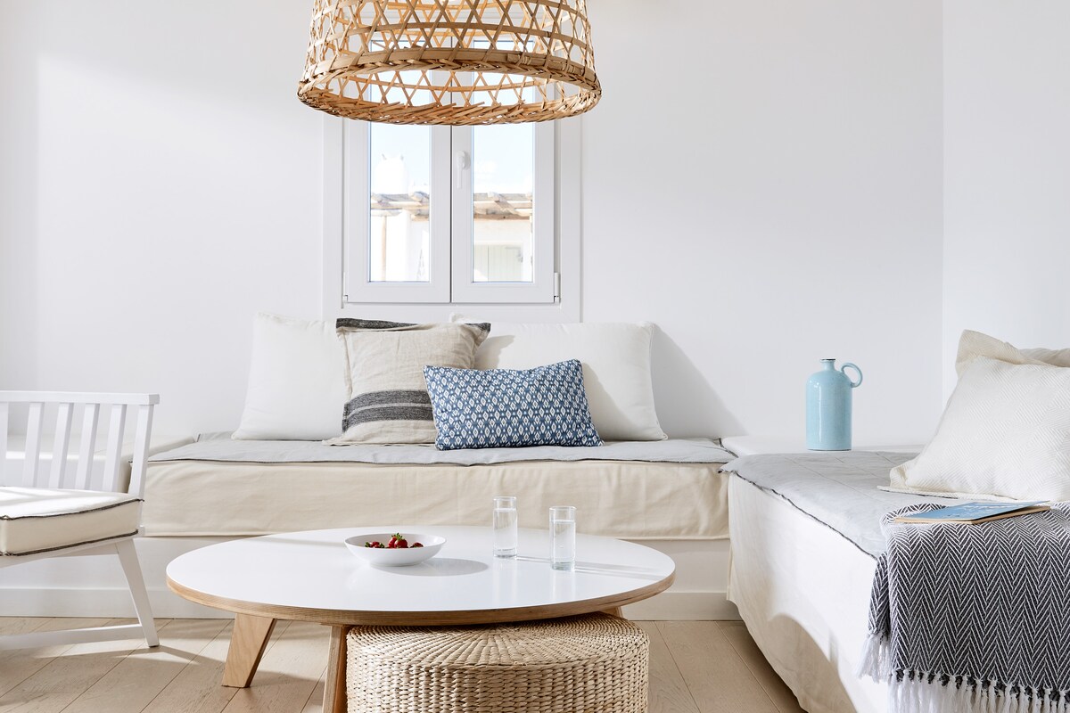 A light-filled living area features a round white table set with a small bowl of fruit. Comfy seating includes a built-in daybed adorned with neutral cushions and a woven pendant light overhead. A white chair is positioned nearby, with a soft throw draped over the edge.