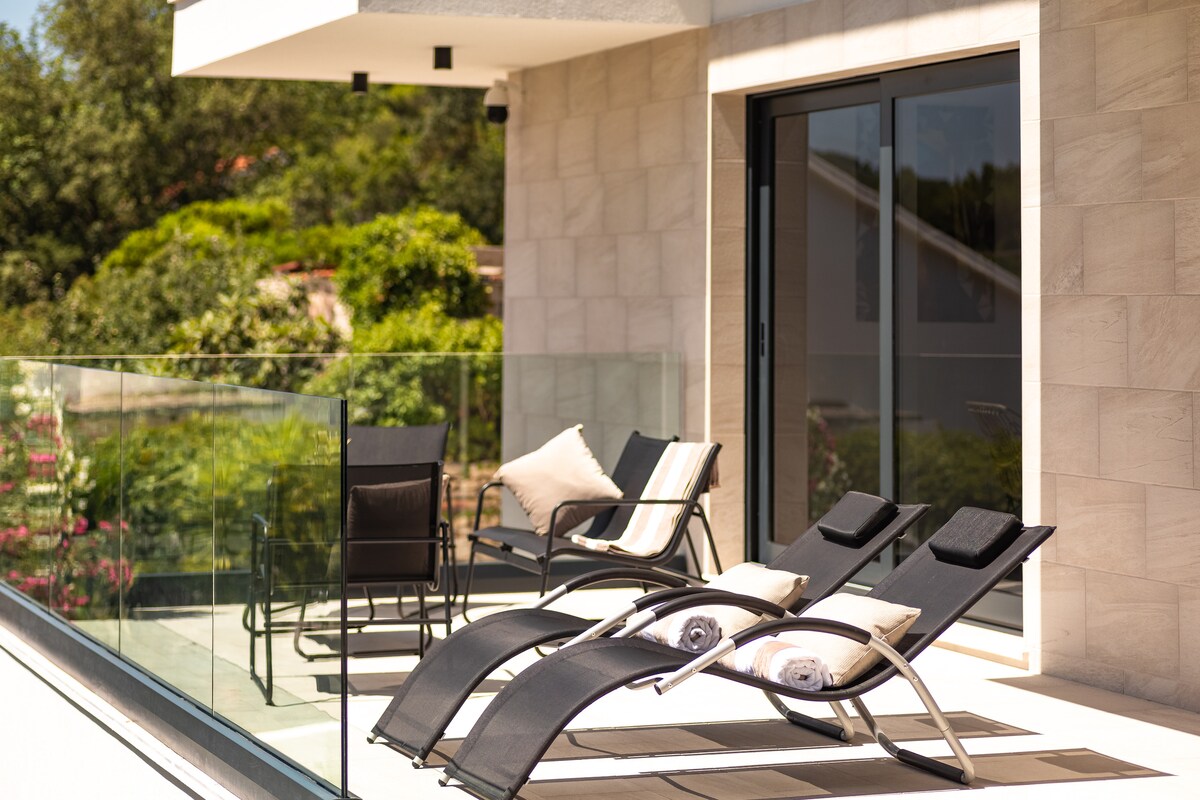 A spacious balcony is furnished with two lounge chairs and a small dining set. Soft cushions are placed on the chairs, and a view of greenery can be seen in the background. The glass railing frames the area, allowing natural light to enhance the space.