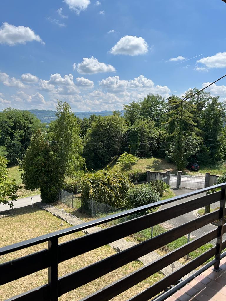 A scenic view from the balcony reveals lush trees and gentle hills under a blue sky dotted with clouds. The nearby road is partially visible, with a glimpse of a vehicle at a distance, creating a serene natural setting.