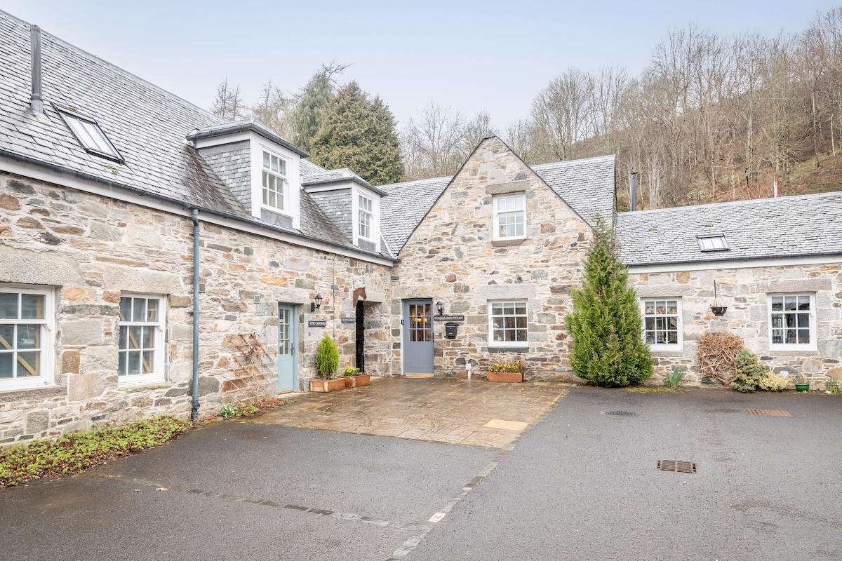 The exterior of a stone cottage is featured, showcasing a charming courtyard with slate-gray roofs and multiple windows. Lush greenery and two small conifer trees are positioned near the entrance. The courtyard is paved, providing direct access to the cottage from the parking area.