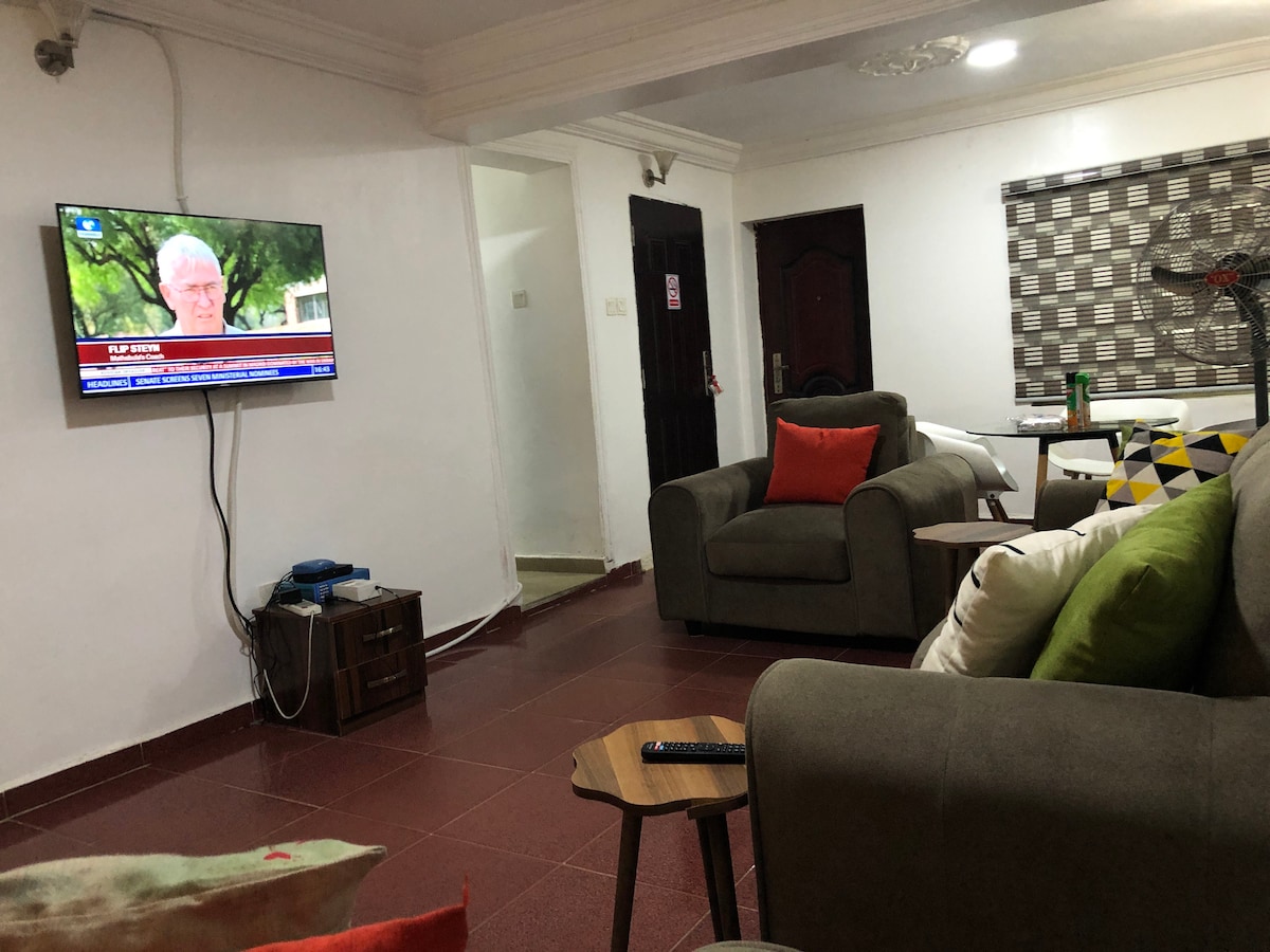 A comfortable living area features a flat-screen television mounted on the wall, with a seating arrangement of an olive green sofa and a patterned armchair. A wooden side table is positioned beside the seating, and a large window is dressed with a geometric privacy curtain.