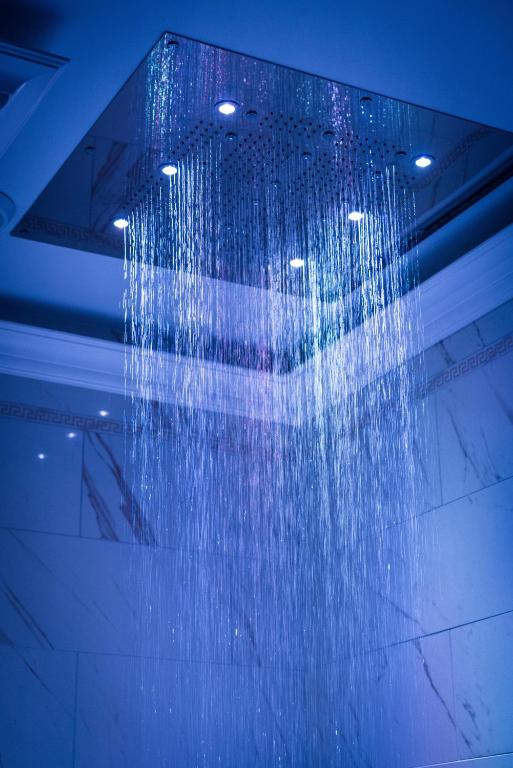 A luxurious rain shower head is featured in an elegant bathroom, with water cascading down in a gentle flow. Soft blue lighting illuminates the shower area, highlighting the glossy marble walls adorned with subtle decorative details.
