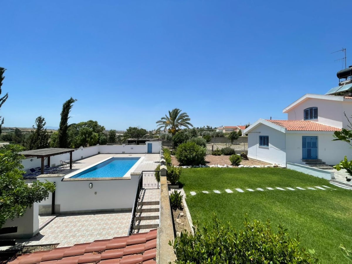 A private pool is set against a backdrop of lush greenery and a clear blue sky. The surrounding garden features well-maintained grass and pathways that lead to a spacious villa. A shaded area with a pergola is visible near the pool, enhancing the outdoor space.