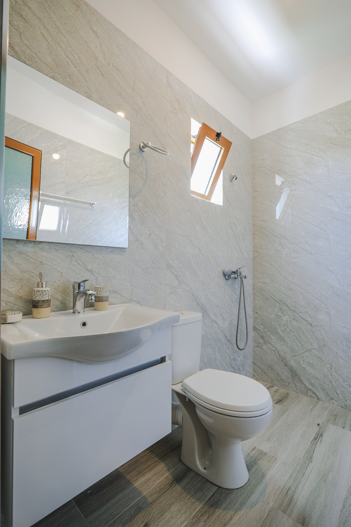 The functional bathroom features a shower cubicle with a glass door, a modern sink with storage underneath, and a large mirror. Natural light from a small window illuminates the space, which is adorned with neutral-toned tiles and a towel rack.