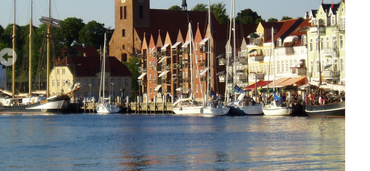 A scenic waterfront view is presented, featuring a row of boats docked along the pier. Colorful buildings with varying architectural styles line the shore, adorned with balconies. In the background, a historic clock tower stands prominently among lush greenery, under a clear blue sky.