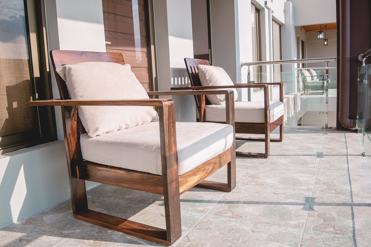 Two spacious armchairs are positioned on a balcony, featuring natural wood frames and soft cushions. The surface is laid with light-colored tiles, and large glass doors provide access to the indoor living space.