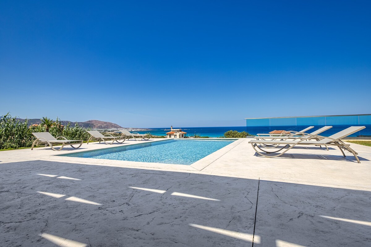 A private pool with a clear blue surface is surrounded by sun loungers on a patio. The view encompasses the turquoise waters of the sea in the background, framed by a clear, cloudless sky. The landscape includes hints of greenery along the edges.
