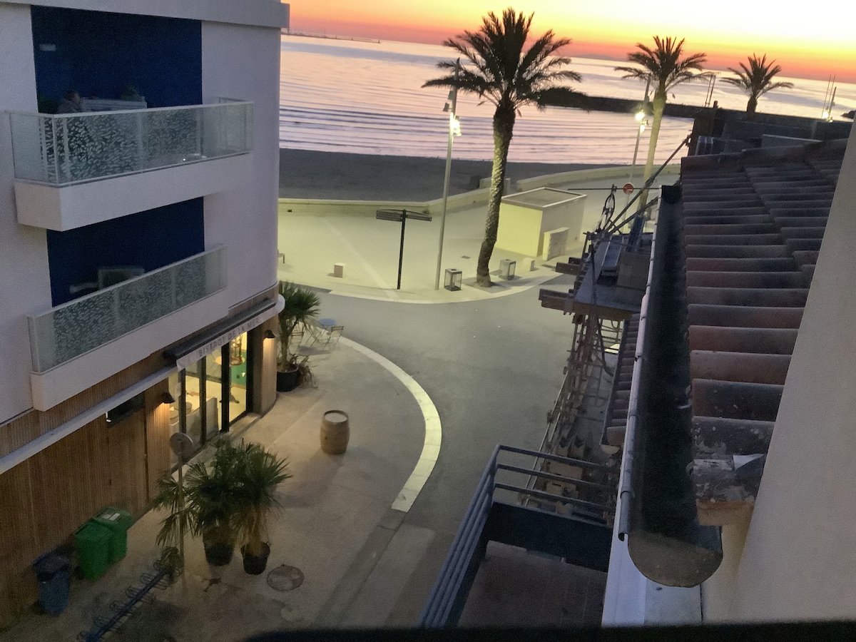 A view of the coastline is presented, featuring a peaceful sunset over the water. Palms are visible along the promenade, and a few buildings are seen, including one with a balcony. The street curves away from the beach, creating a tranquil atmosphere.