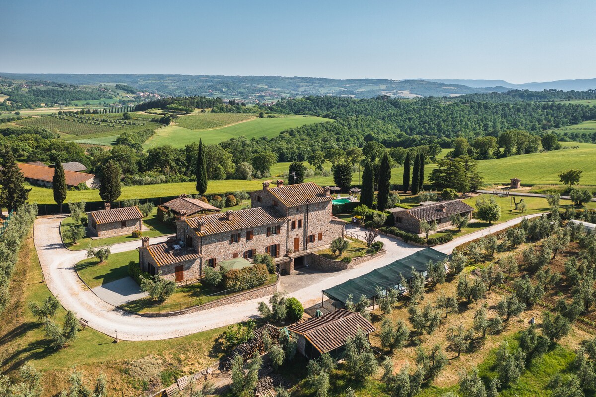 An aerial view of a rustic farmhouse compound, surrounded by lush green hills and olive groves. Several cottages and a main building are displayed, along with parking spaces and pathways leading into the countryside. The expansive rural landscape extends into the distance under clear blue skies.