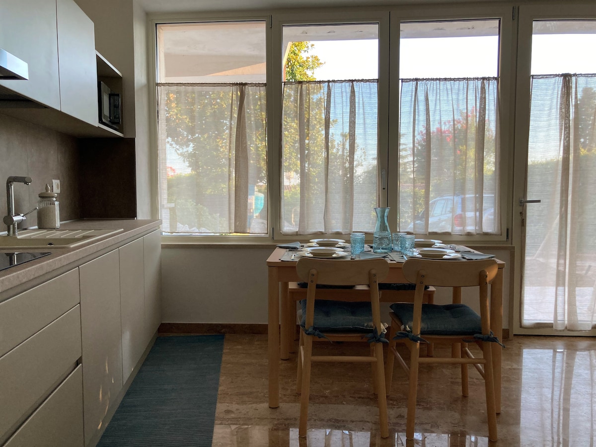 A modern kitchen area features large windows with light curtains, allowing natural light to brighten the space. A simple wooden dining table is set for four, accompanied by light blue placemats. Neutral cabinetry and a countertop are present, providing ample workspace.