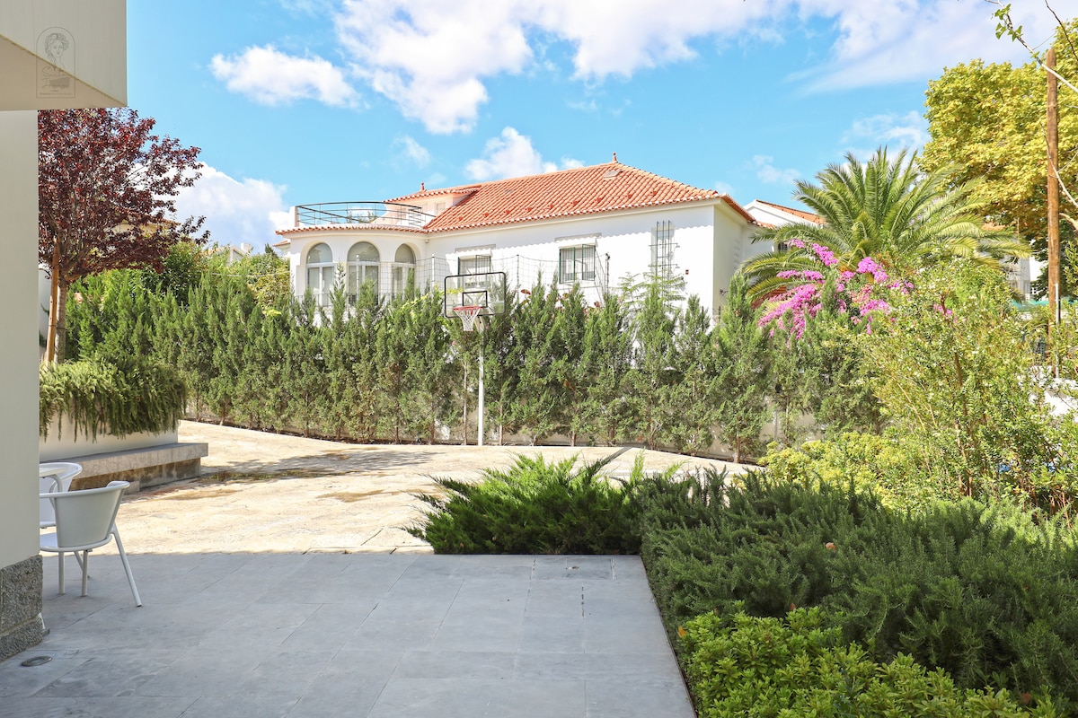 A serene outdoor space provides a clear view of lush green hedges and palm trees surrounding a white villa. The bright sky is partially covered by clouds, indicating a pleasant day. A patio area with simple seating is visible, setting a tranquil atmosphere.