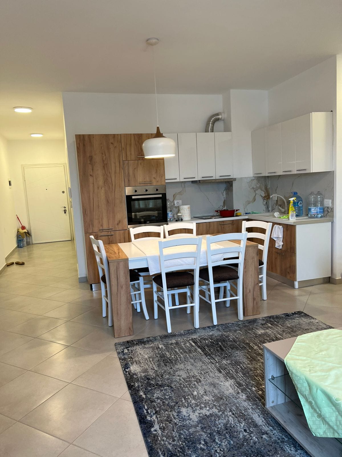 A modern kitchen and dining area are shown, featuring a wooden dining table with white chairs. The kitchen includes contemporary cabinetry and appliances, and light tiles cover the floor. A soft area rug adds comfort to the space.