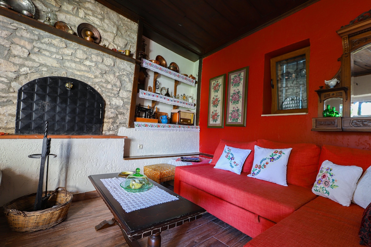A cozy living area features a bright red wall providing a vibrant backdrop for a comfortable sofa adorned with floral cushions. A stone fireplace is prominently placed, and shelves display various decor items. Natural light filters through a small window, adding warmth to the space.