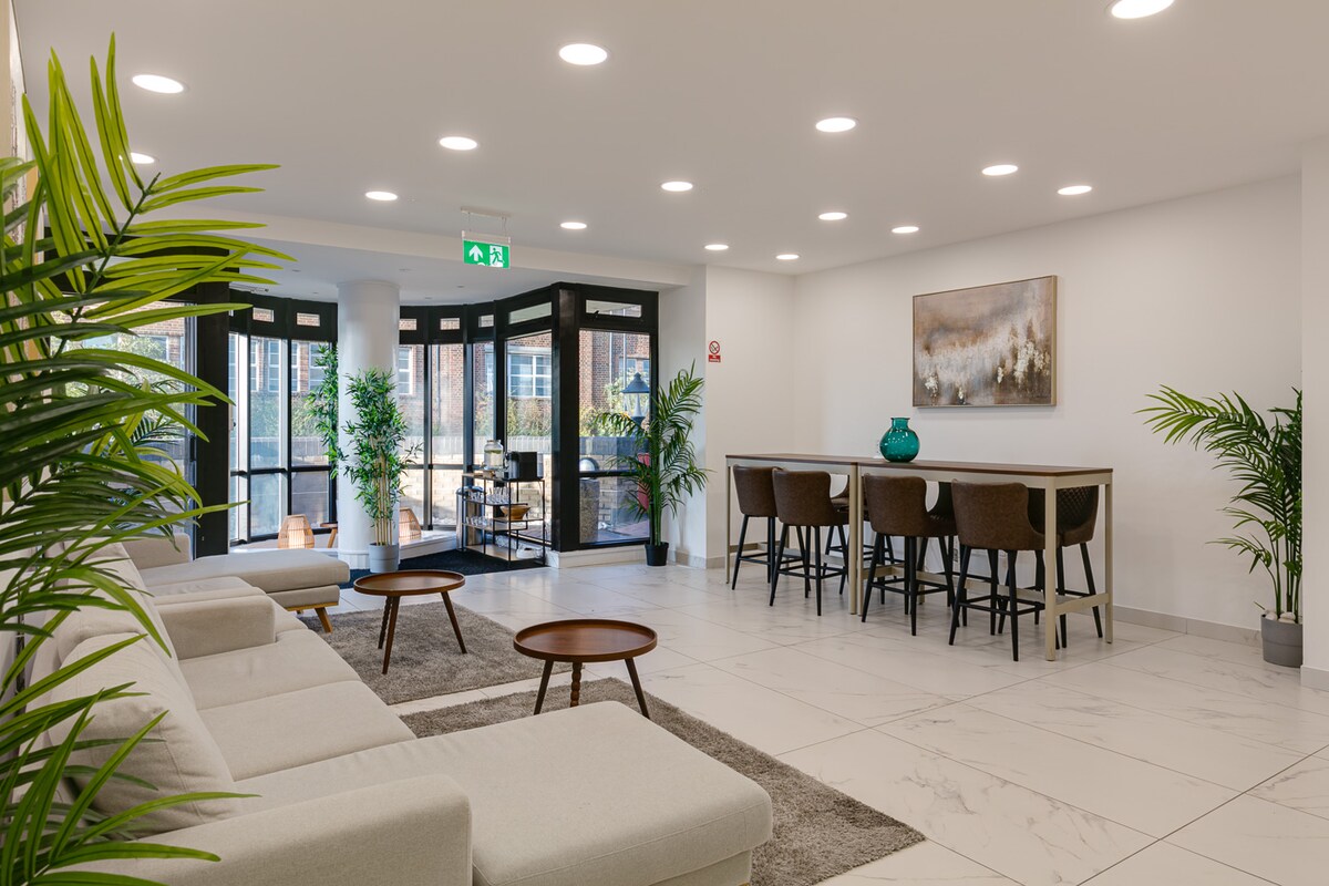 A modern lobby area features a light-filled space with a comfortable seating arrangement, complemented by greenery. Two round tables sit alongside a long dining table with high chairs. Neutral tones are present in the decor, providing an open and airy atmosphere.