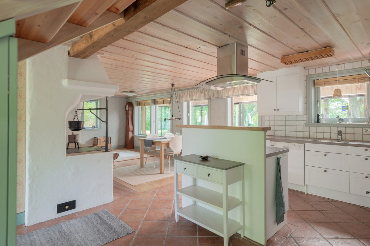 An open-plan kitchen and dining area features wooden beams across the ceiling and terracotta tile flooring. Natural light pours in through multiple windows, illuminating white cabinetry and a central island. A dining table is set for four, complemented by a cozy seating corner in the background.