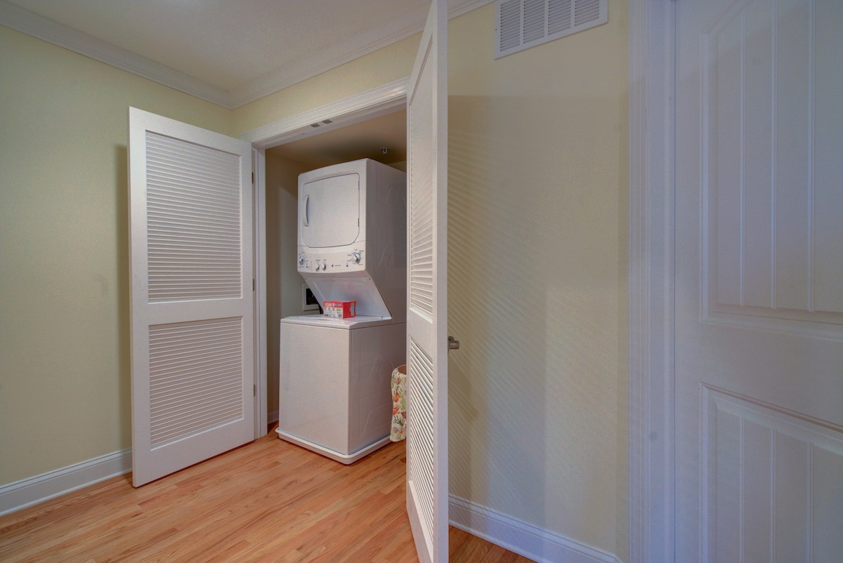 A laundry area is shown with a stacked washer and dryer unit positioned against a neutral wall. Light wood flooring extends throughout the space, while the open doors of the closet reveal the appliances, providing accessibility for household tasks.