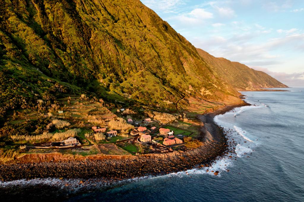 An aerial view captures the scenic coastline of Fajã do Belo, lined with lush green mountains. Several self-catering houses are nestled among the landscape, with rocky shores and gentle waves in the foreground, creating a serene connection between nature and accommodation.