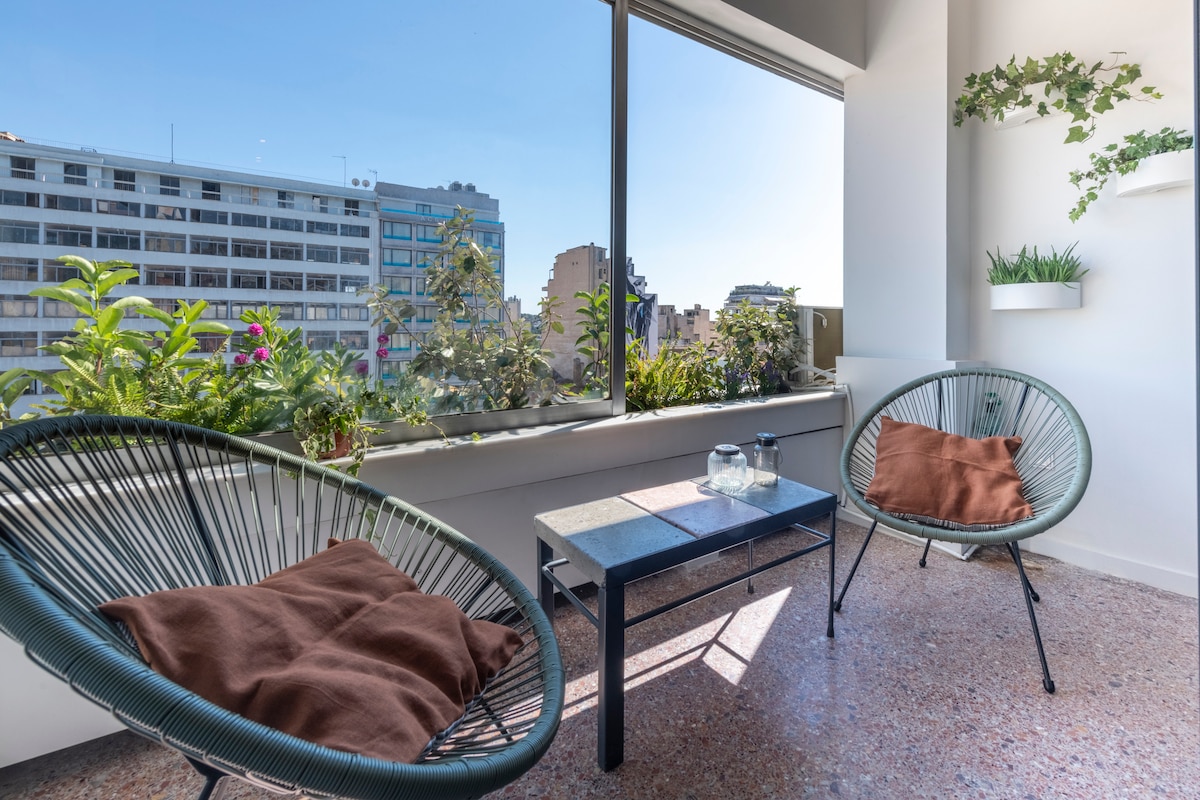 A bright seating area features two woven chairs facing large windows. A small table is positioned between them, with a clear view of the city skyline. Potted plants adorn the window ledge, enhancing the natural light in the space.