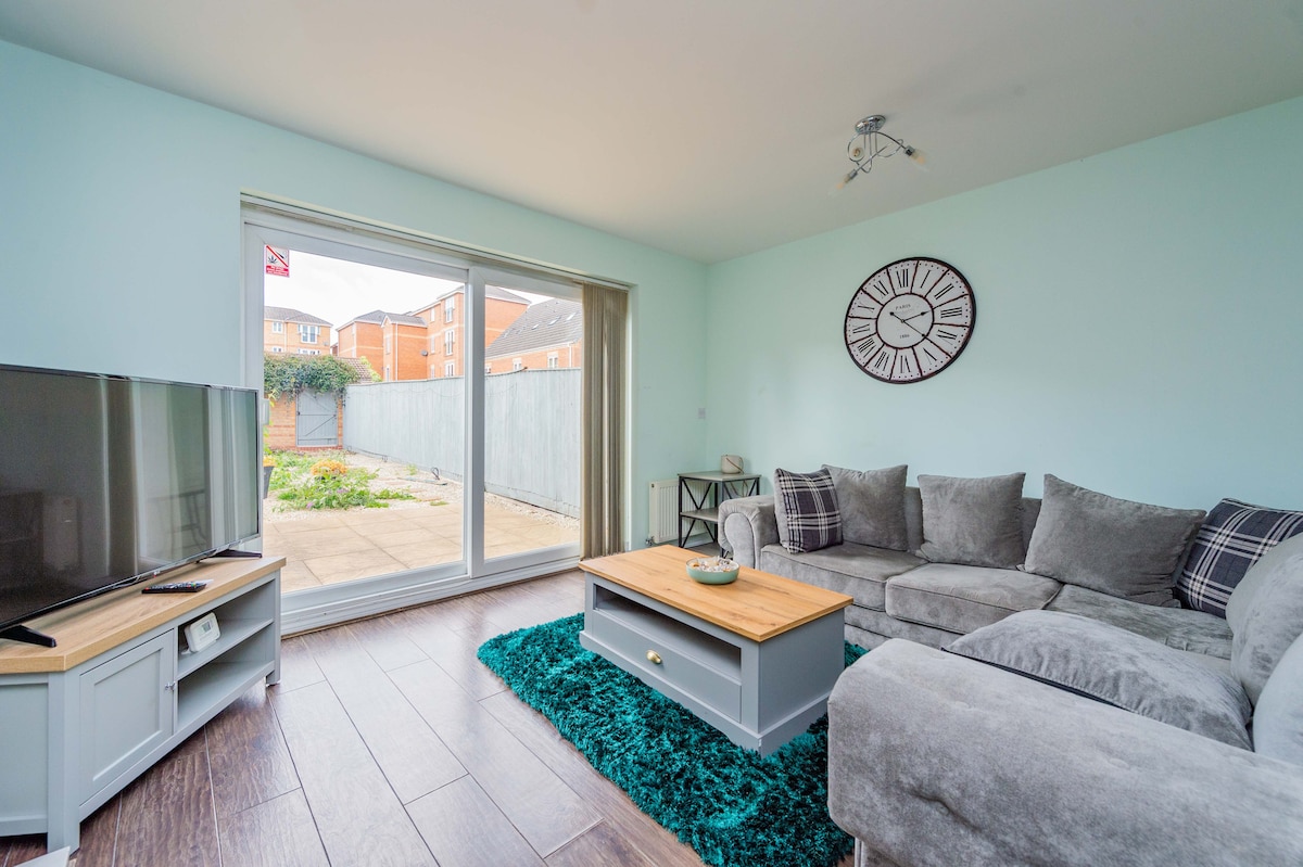 A modern living area features a plush sectional sofa in gray, complemented by a wooden coffee table. A large TV is positioned on a white console. Natural light floods the space through expansive glass doors, which lead to a private garden area, enhancing the sense of openness.