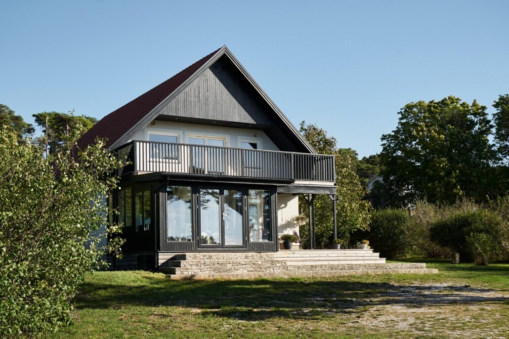 A modern two-story house is showcased, featuring a dark grey wooden exterior and a spacious balcony. Large glass doors provide ample natural light, while a well-maintained lawn surrounds the property. The structure is set against a clear sky, highlighting the tranquil setting.