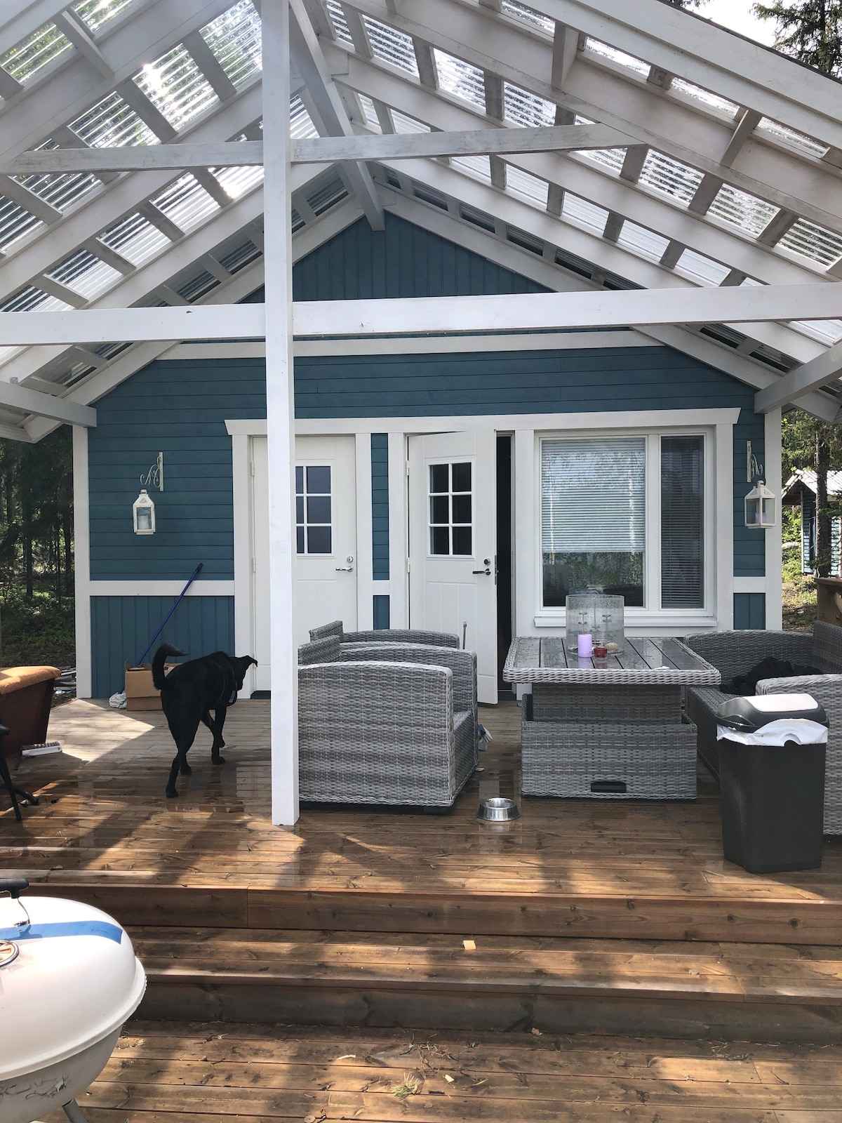 A covered terrace is featured in the front of the cottage, showcasing an outdoor seating area with wicker chairs and a table. Two entrance doors lead into the interior, with a large window allowing natural light to enter. A pet can be seen walking nearby.