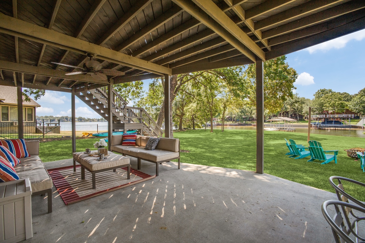 A covered outdoor seating area features a comfortable sectional sofa with decorative cushions. A coffee table sits in the center, surrounded by a striped rug. Lush green lawn extends towards the lake view, framed by mature trees. Stairs lead up to the upper level.