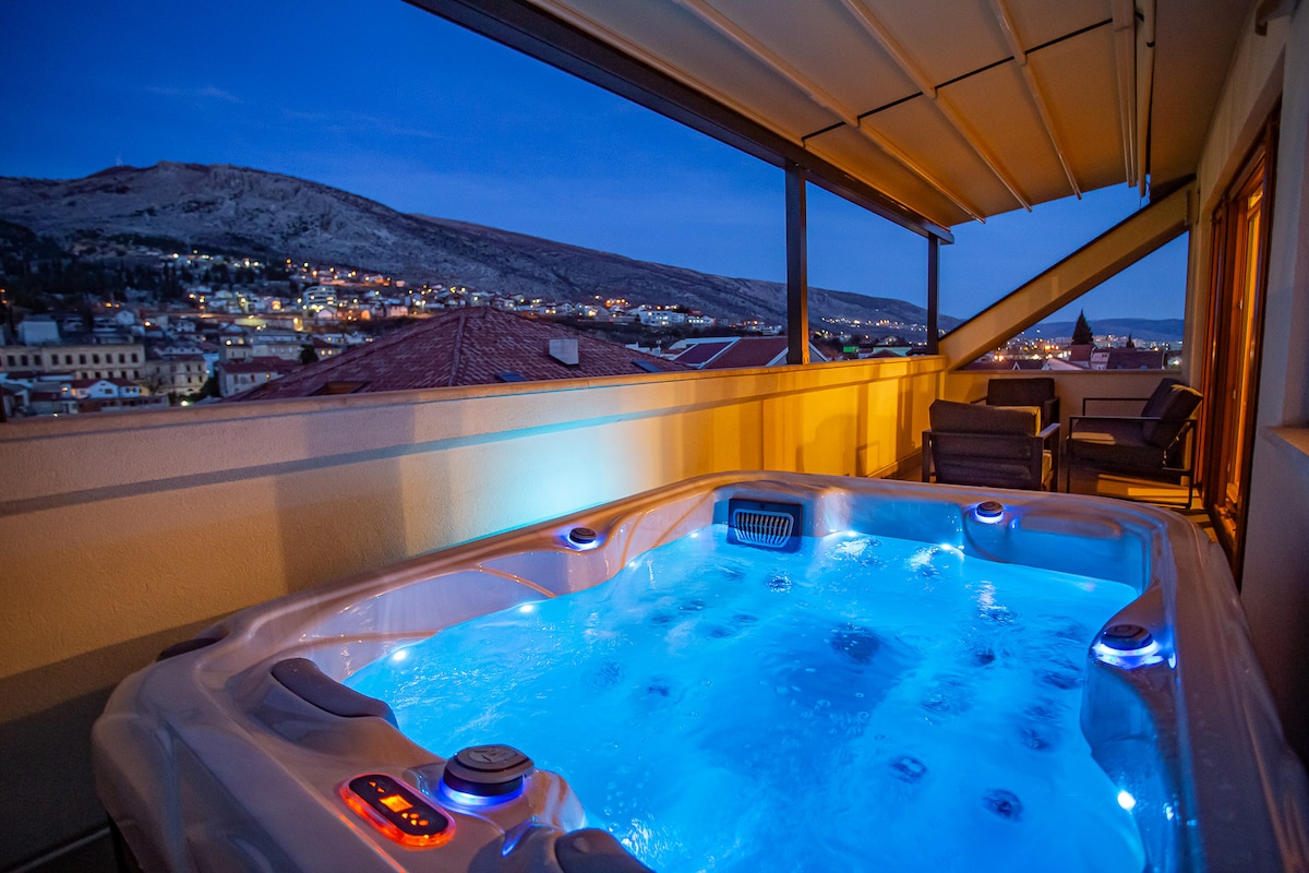 A private hot tub is set on a balcony with panoramic views of the surrounding hills and cityscape. Evening lighting creates a serene atmosphere, highlighting the tub's illuminated water. Comfortable seating is arranged nearby, providing a welcoming space for relaxation.