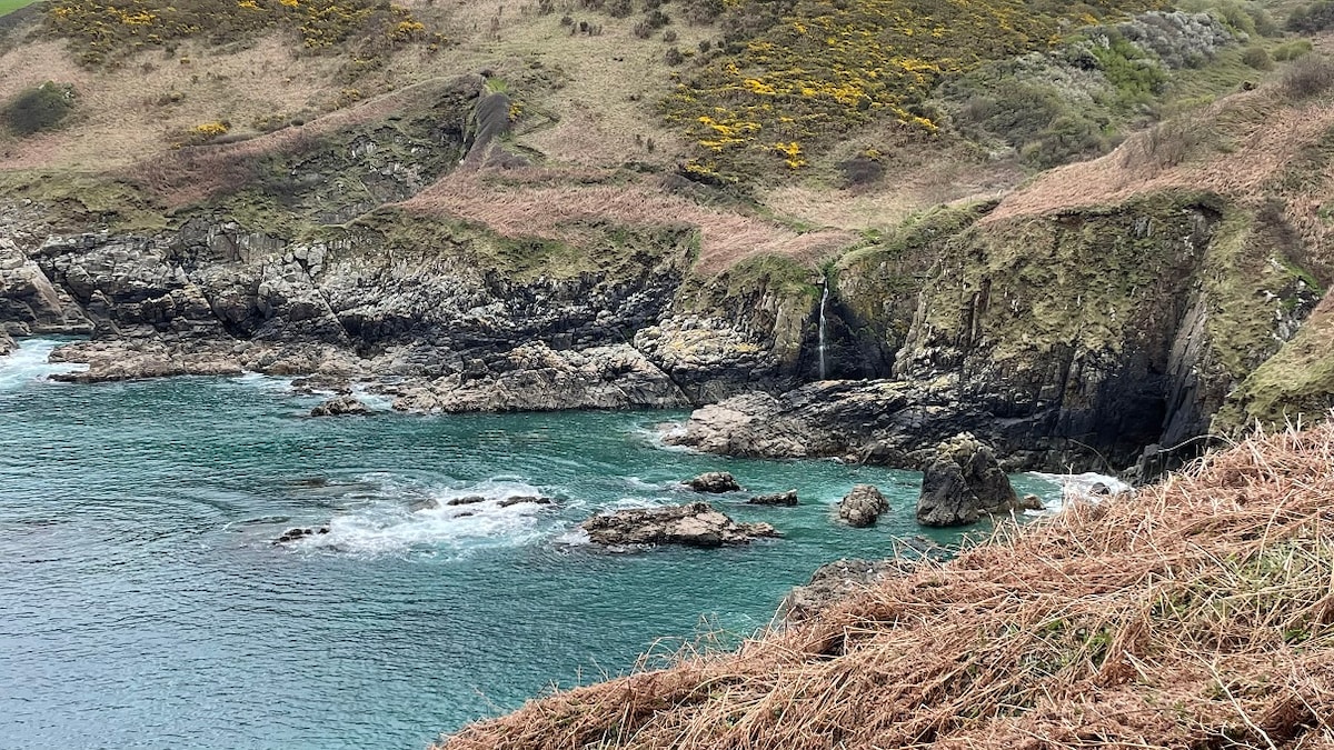 A coastal view showcases rocky cliffs and clear blue waters. Waves gently lap against the shore, while patches of vibrant vegetation peek through the rugged landscape. The terrain rises in the background, creating a scenic backdrop of rolling hills.