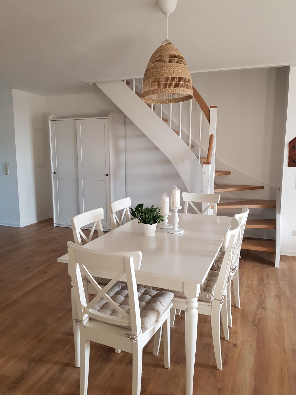 An open dining area features a white table surrounded by six chairs with padded seats. Light wood flooring complements the space. A decorative plant sits in the center of the table, while a stylish, woven pendant light hangs overhead. A staircase leads to the upper level.