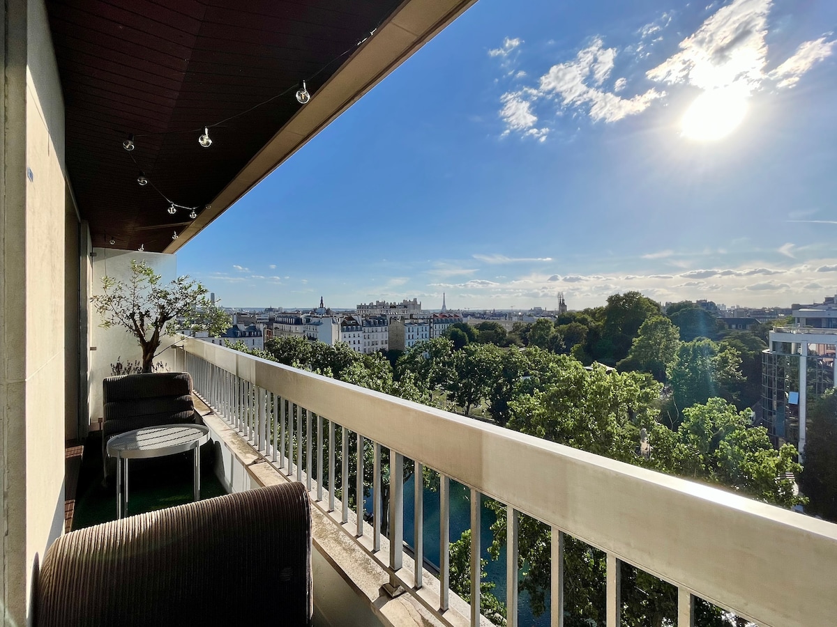 A spacious balcony overlooks a vibrant cityscape, showcasing greenery and urban architecture. Comfortable seating arrangements and outdoor lighting create a welcoming space. The clear sky allows for ample natural light, enhancing the panoramic view of Paris.