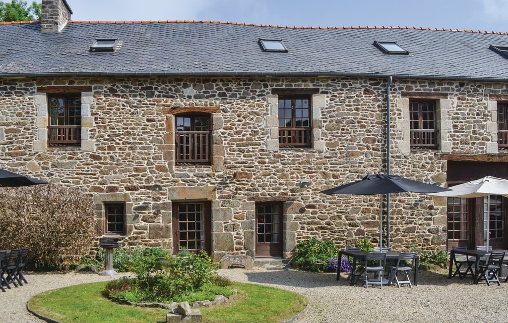 A historic stone building is showcased with multiple windows framed in wood. The facade is complemented by outdoor seating arrangements under large umbrellas and a variety of greenery, enhancing the inviting courtyard space.
