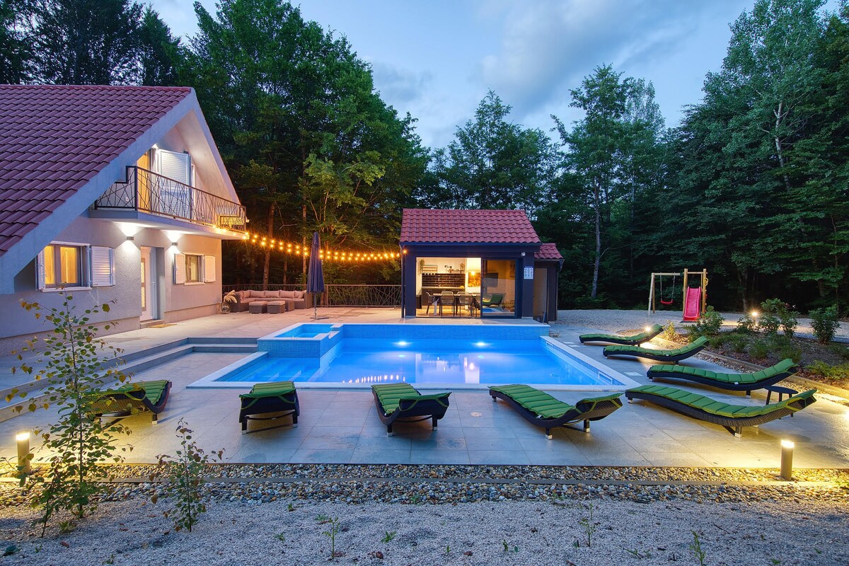 A serene outdoor space is highlighted by a rectangular swimming pool surrounded by lounge chairs. Nearby, a cozy seating area is illuminated by string lights. The two-story house features a red-tiled roof and large glass doors that open to the patio.